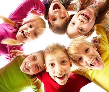 A circle of smiling children with linked arms leans into the center of the circle to look down at the camera.